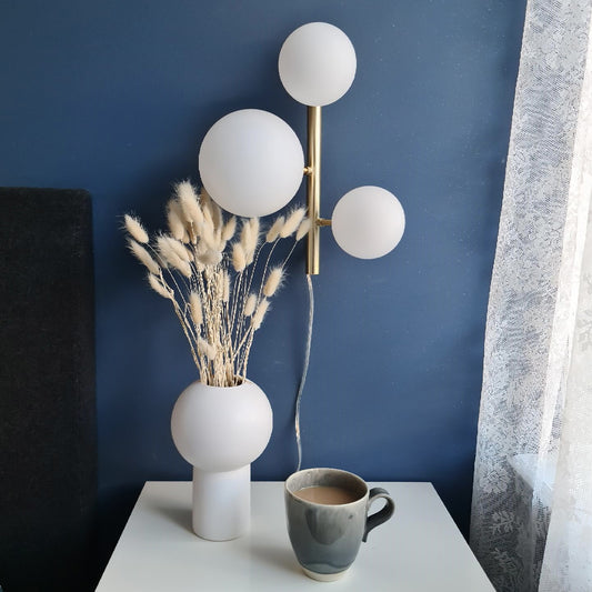 Molekyl vegglampe som sengelampe på et soverom, laget i messing metall og tre opale glasskupler festet langs en loddrett stang - Aneta belysning