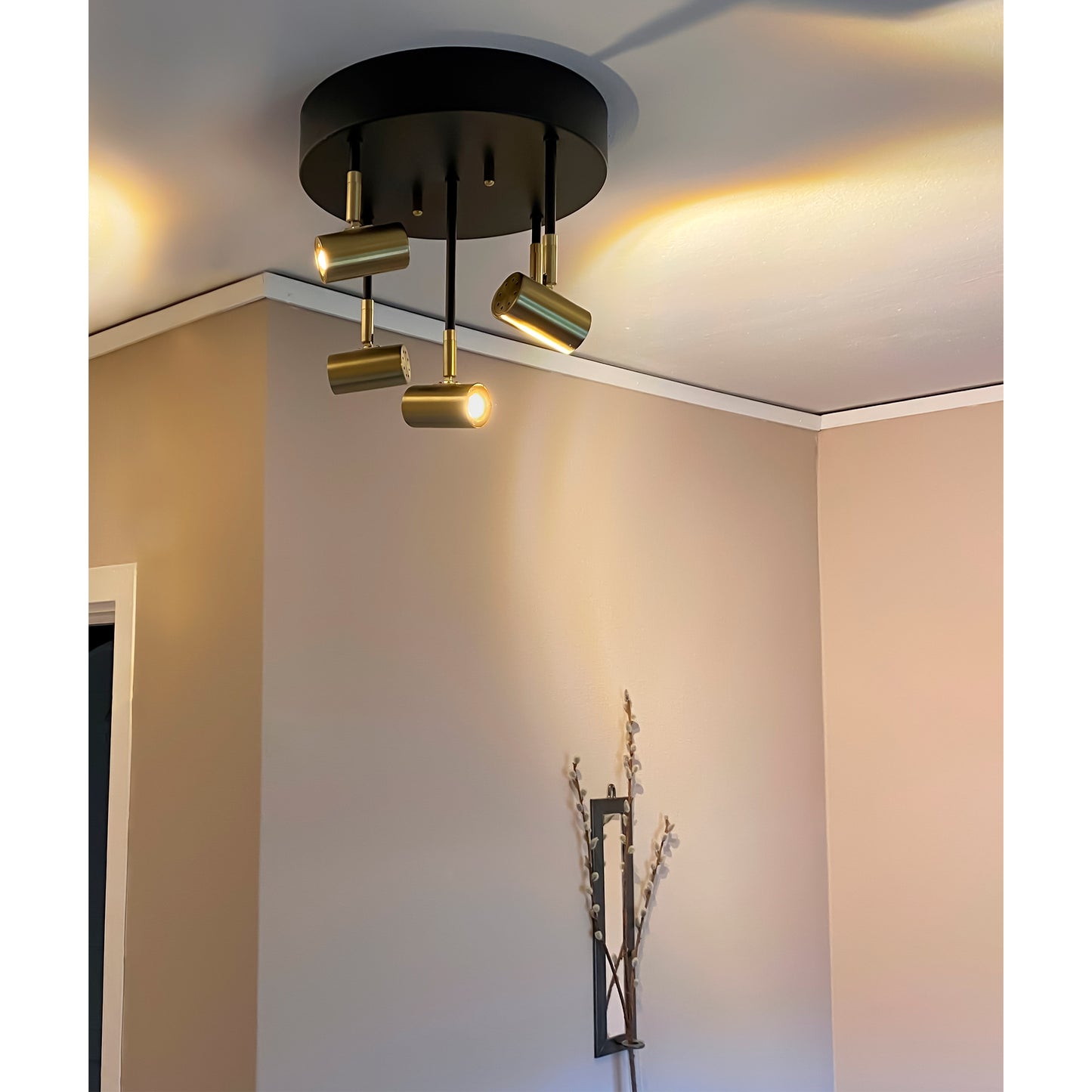Gusto spotlight taklampe lysende i en gang med fem justerbare spotter i ulik høyde plassert på en rund takplate, laget i sort metall og messing - Aneta belysning