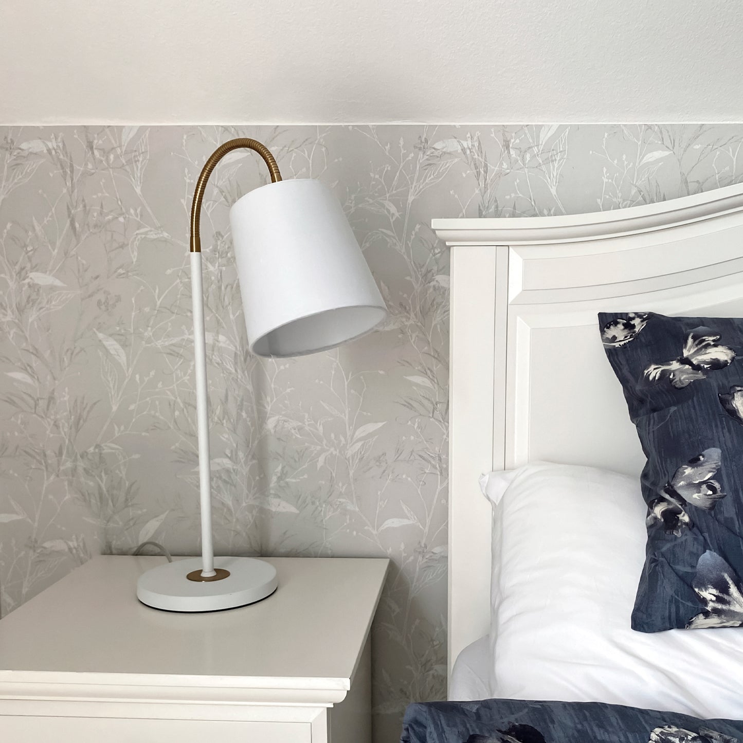 Ljusdal bordlampe plassert på en nattbord på soverom. Laget i hvit metall med ledd i messing og hvit lampeskjerm - Aneta belysning