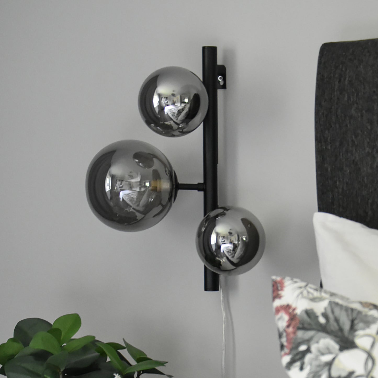 Molekyl vegglampe som nattbordslampe på et soverom, laget i sort metall og tre sotede glasskupler festet langs en loddrett stang - Aneta belysning